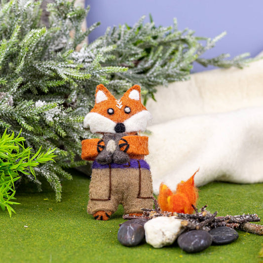 A handcrafted felt fox ornament dressed in  brown pants and holding a pair of binoculars, standing beside a small felt campfire on artificial green grass, with frosted greenery and a soft cream fabric backdrop.