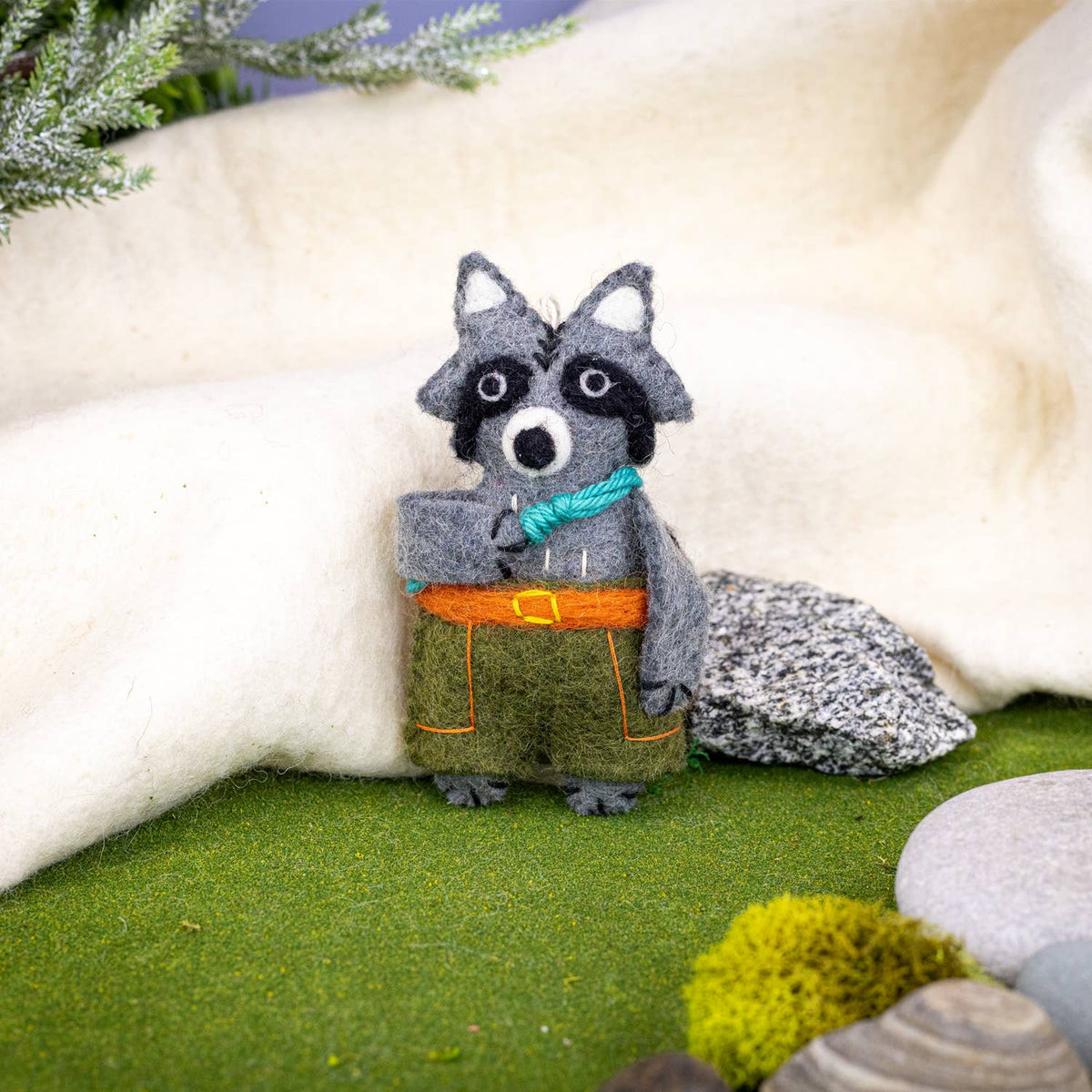 Felted raccoon ornament with a blue climbing rope and green pants in a natural setting