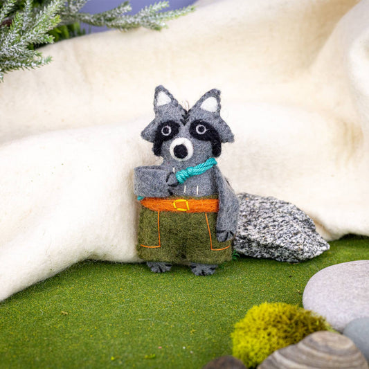 Felted raccoon ornament with a blue climbing rope and green pants in a natural setting
