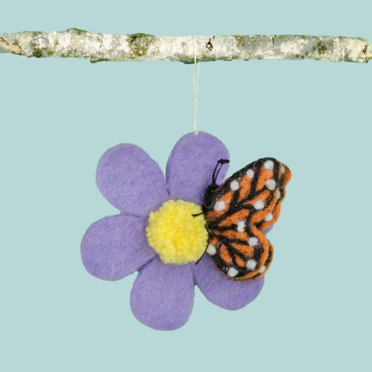 A monarch butterfly rests on the yellow pom-pom center of a purple flower. Hung from a birch branch by a white string against a blue background.