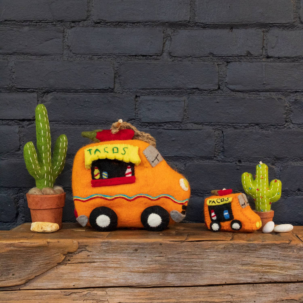 A handmade felt taco truck decoration in orange and yellow sits on a rustic wooden surface against a dark gray brick wall, styled with small potted cacti.