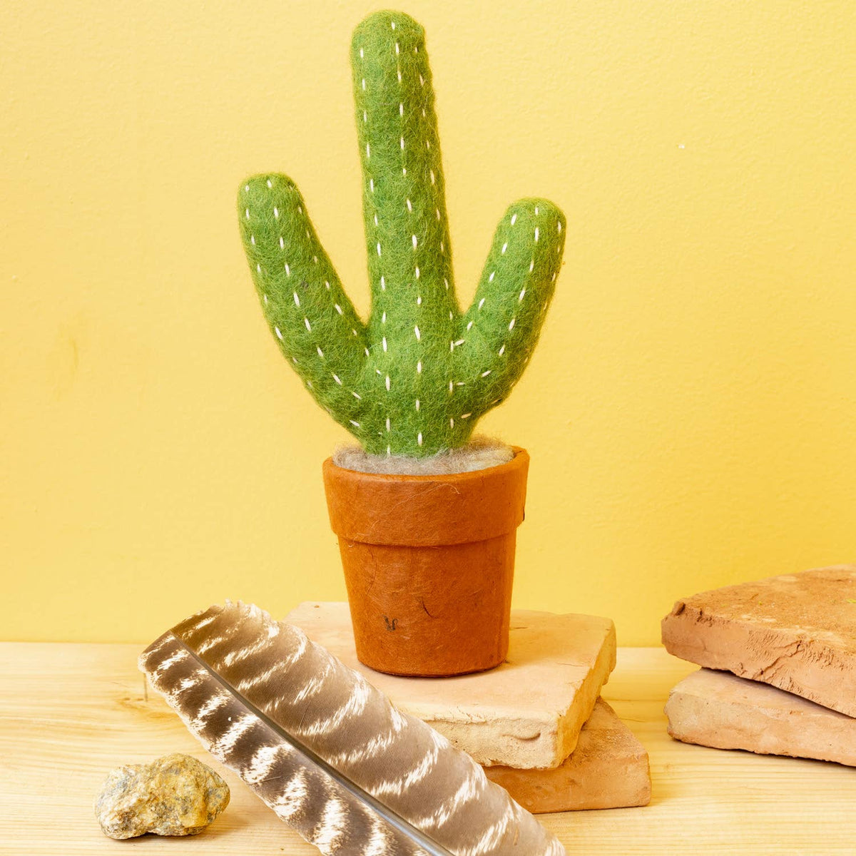 A green felt cactus in a terracotta pot, styled on a wooden surface with a feather, stones, and stacked tiles against a yellow background.
