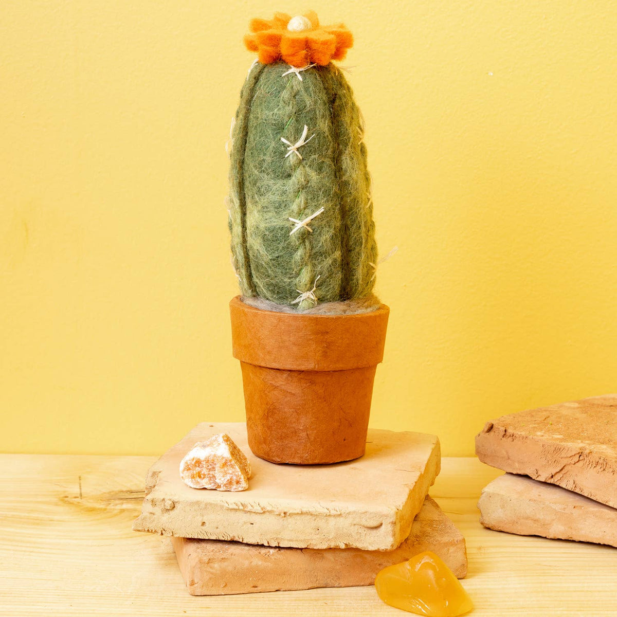 A felted wool cactus in a terracotta pot, styled on stacked stone tiles against a solid yellow background.