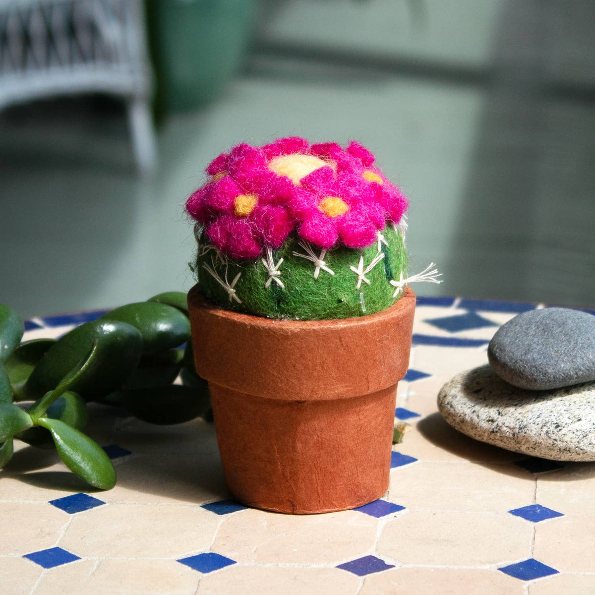 A felt cactus plant in a terracotta-colored pot, decorated with bright pink flowers and green felt foliage, set on a tiled surface with decorative stones and a blurred background.