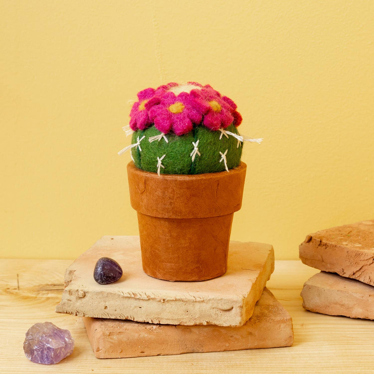 A small felt cactus with bright pink flowers and yellow centers, planted in a terracotta-colored pot, displayed on stacked stone slabs against a warm yellow background.