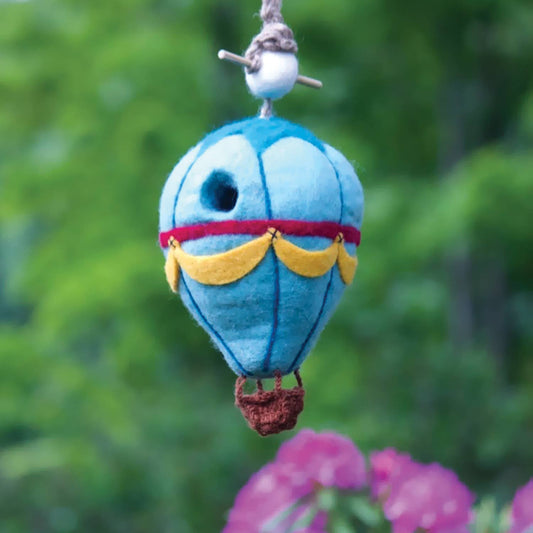 A small felt hot air balloon bird house hanging outdoors against a blurred green garden background with pink flowers in the foreground. The balloon is light blue with a red band, yellow scalloped trim, a brown basket, and a white bead with gray string at the top.