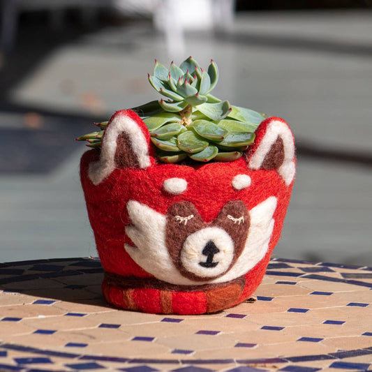 A red felt planter shaped like a red panda head, holding a green succulent, placed on a tiled surface in an outdoor setting.