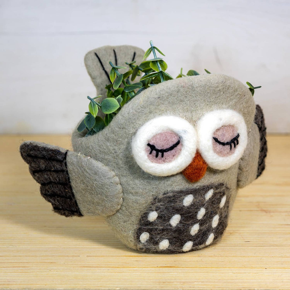A gray felt owl planter with embroidered closed eyes, a small orange beak, dark gray wings, and a white polka-dotted belly, holding green leaves and set on a light wood surface against a white wall.