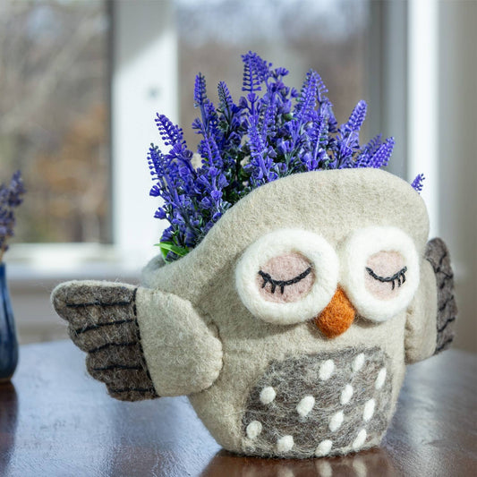 A felt owl-shaped planter in light gray with embroidered closed eyes, an orange beak, and white polka dots on the belly, holding purple flowers, set on a wooden table near a window.