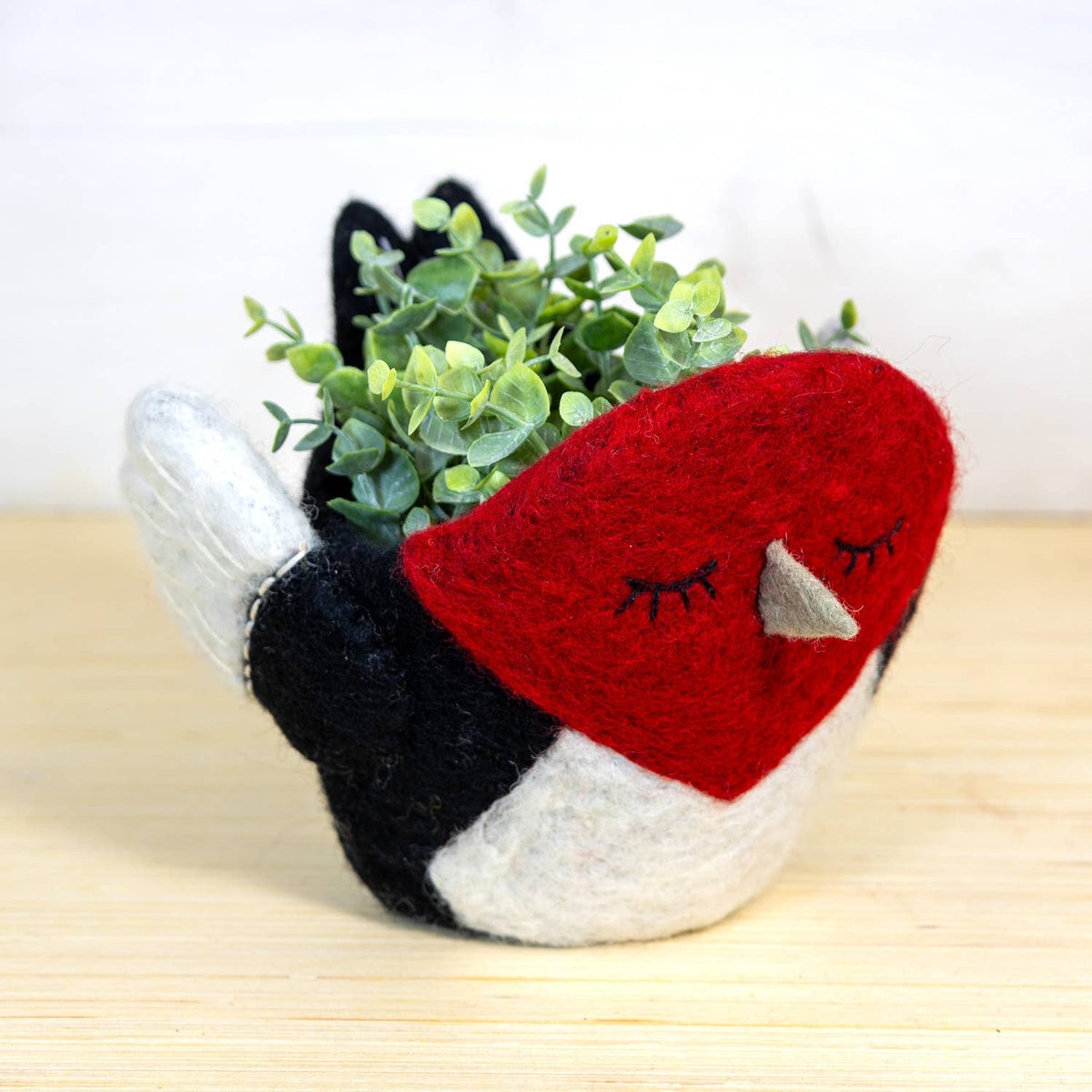 A felt bird-shaped planter with a red heart-shaped face, black and white body, and small beak, holding green eucalyptus stems, set on a light wood surface against a white background.