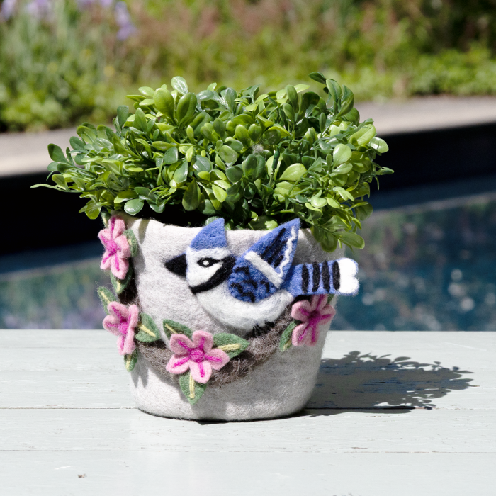 A felt planter pot with a blue jay decoration and pink flowers, holding a green leafy plant, set on a white wooden surface with a blurred outdoor background.