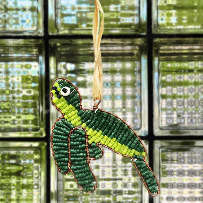 A green sea turtle shaped ornament made with glass beads and wire hanging by a piece of raffia sitting on a background of green glass tiles.