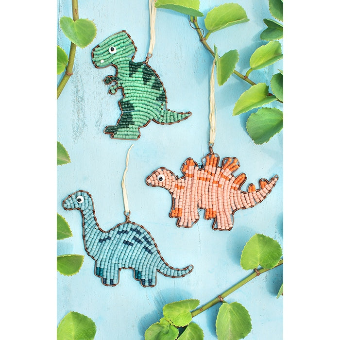 Three beaded dinosaur ornaments hanging from white strings against a light blue background with green leaves around the edges. The dinosaurs are colored green, blue, and orange.