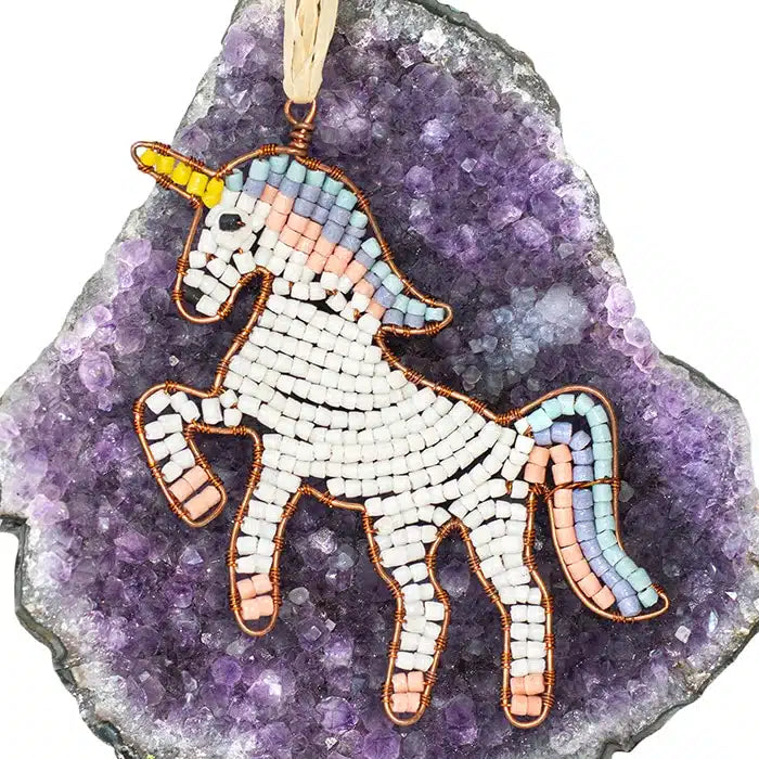 a unicorn shaped ornament hangs by a piece of raffia. The ornament is make from individual recycled glass beads intertwined with copper wire, sitting on a large purple amethyst. 