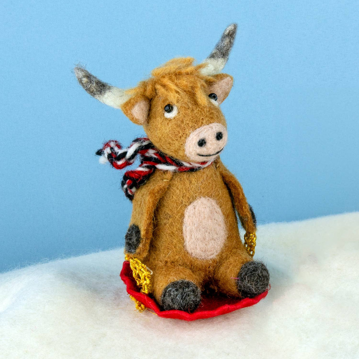 Felted toy bull ornament with a red sled on a white surface with blue background
