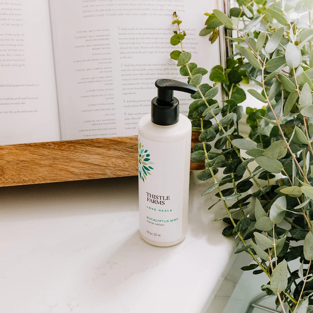 Thistle Farms eucalyptus mint hand lotion bottle on a surface with a plant and book in the background.