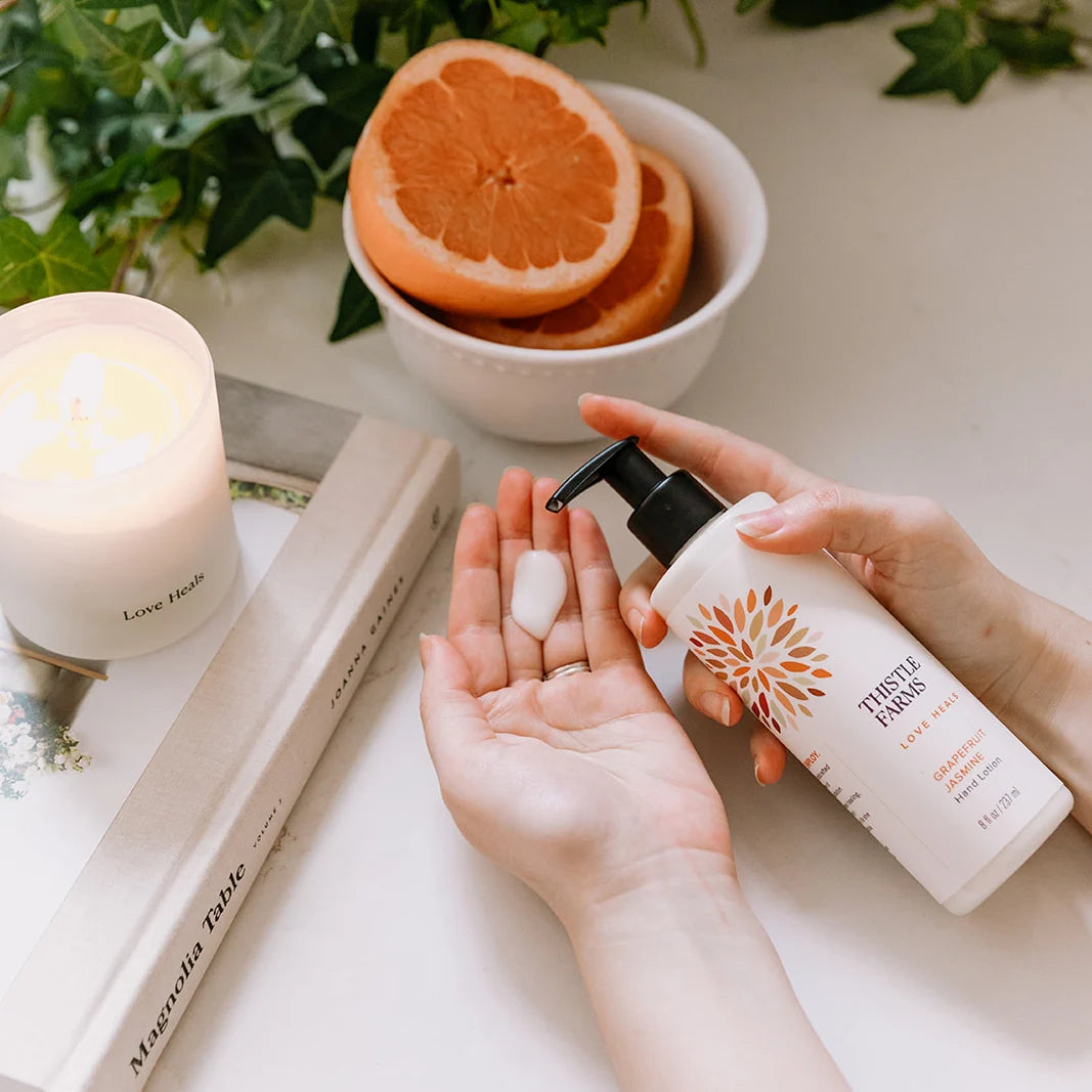 Person applying lotion from a bottle labeled 'Thistle Farms' with a bowl of grapefruit and a candle in the background.