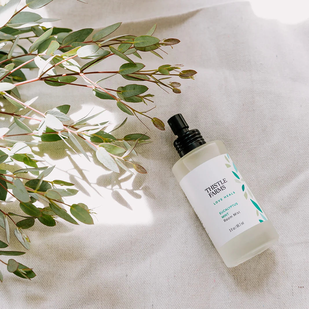 Thistle Farms bottle of eucalyptus room mist on a textured surface with green leaves.