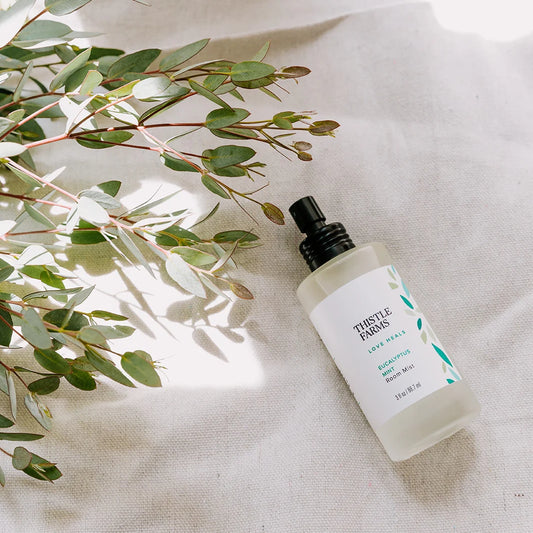 Thistle Farms bottle of eucalyptus room mist on a textured surface with green leaves.