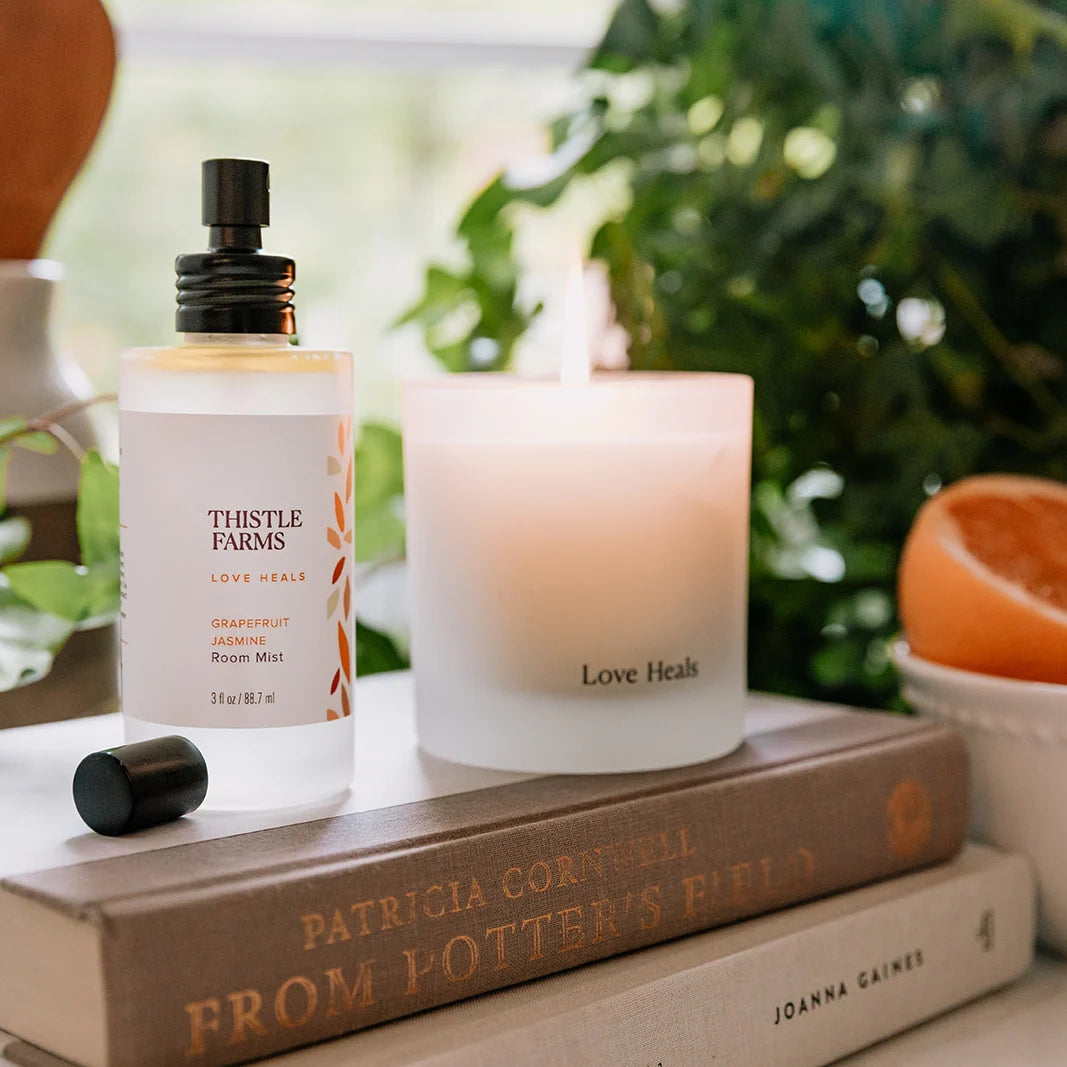 Thistle Farms room mist bottle and candle on books with a blurred green background