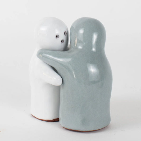 Two glossy ceramic figurines shaped like abstract human figures embracing, one white with small dark eye details and one pale blue-gray, on a clean white background.