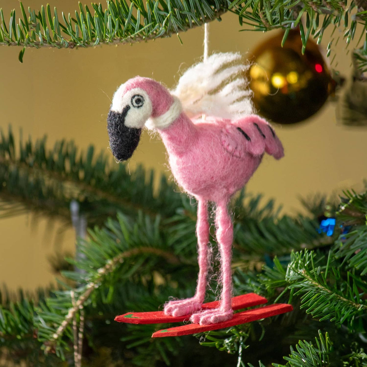 A pink felted wool flamingo ornament hanging from a green Christmas tree branch, with red wooden skis underneath and a gold ornament blurred in the background.