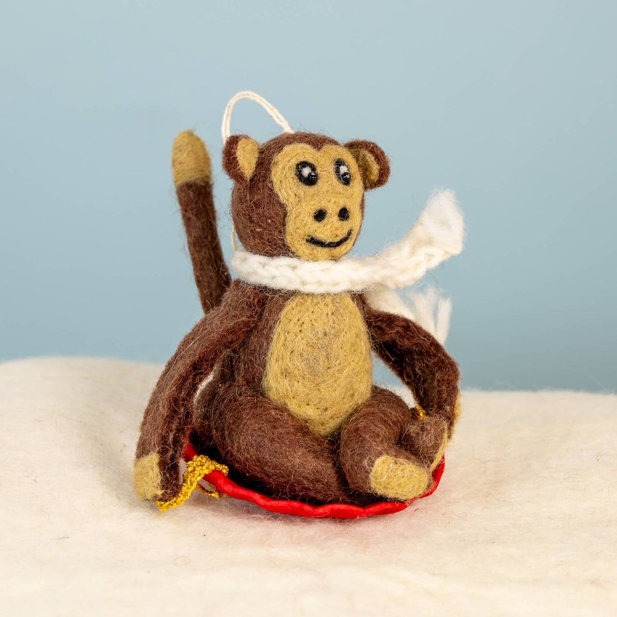 A felt monkey ornament sitting on a red sled with a white knit scarf, photographed against a light blue background and white surface.