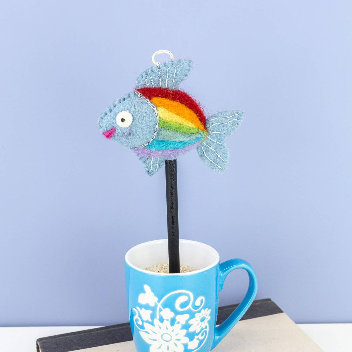 A felt fish-shaped pencil topper with a rainbow stripe pattern and a silver hanging loop, shown perched on top of a blue mug against a light blue background.