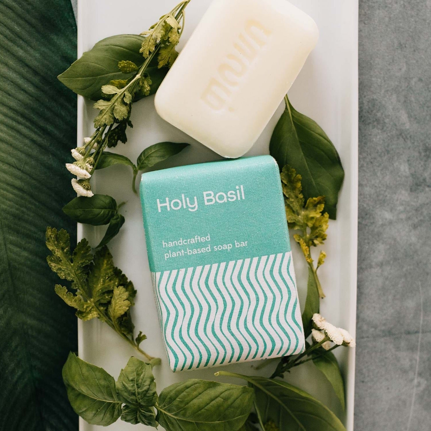 A white bar of soap and its teal and white rectangular package are laying on a white tray and surrounded by green leaves and small flowers.