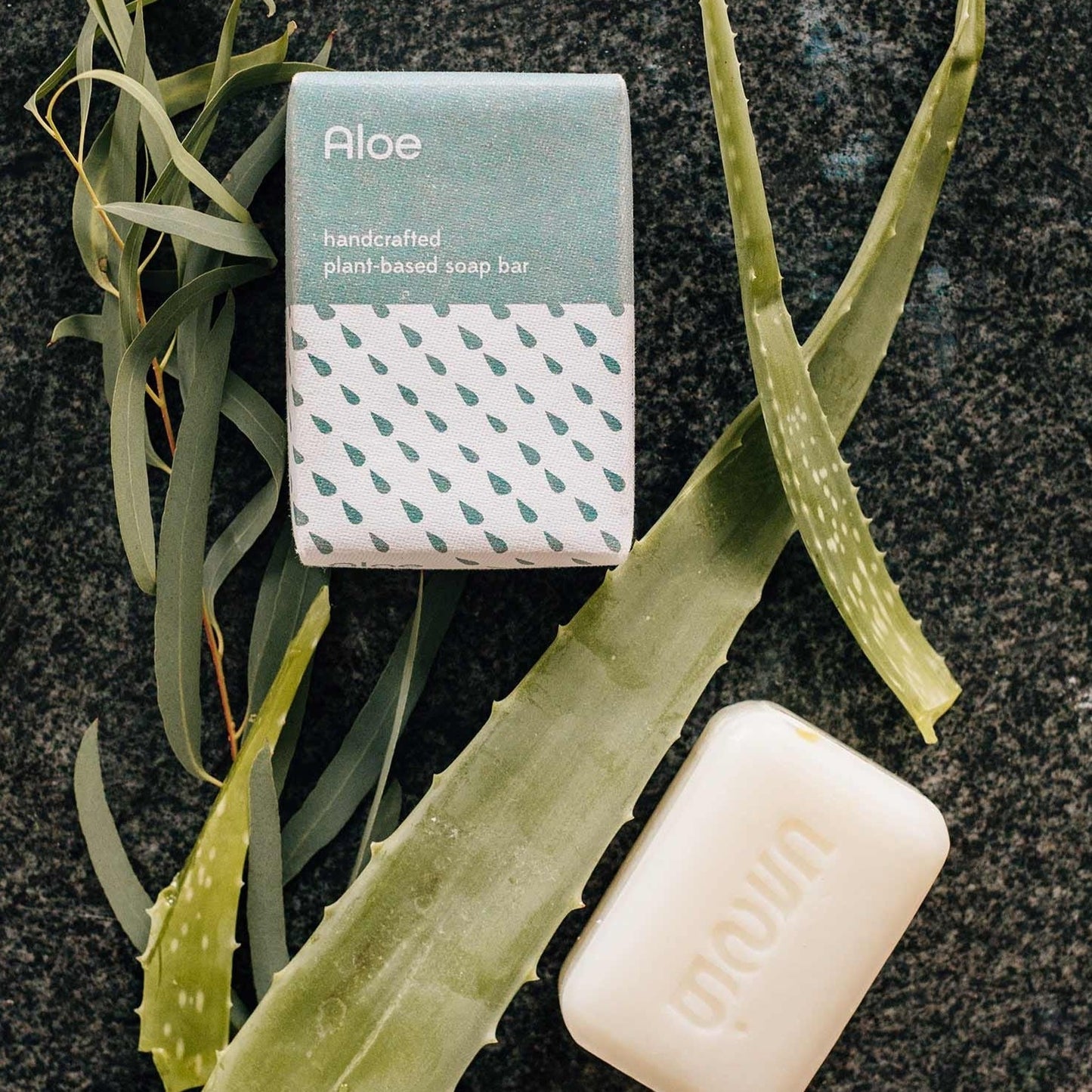 A bar of white aloe scented soap and it's blue and white packaging is placed on a black stone background and shown with aloe leaves.