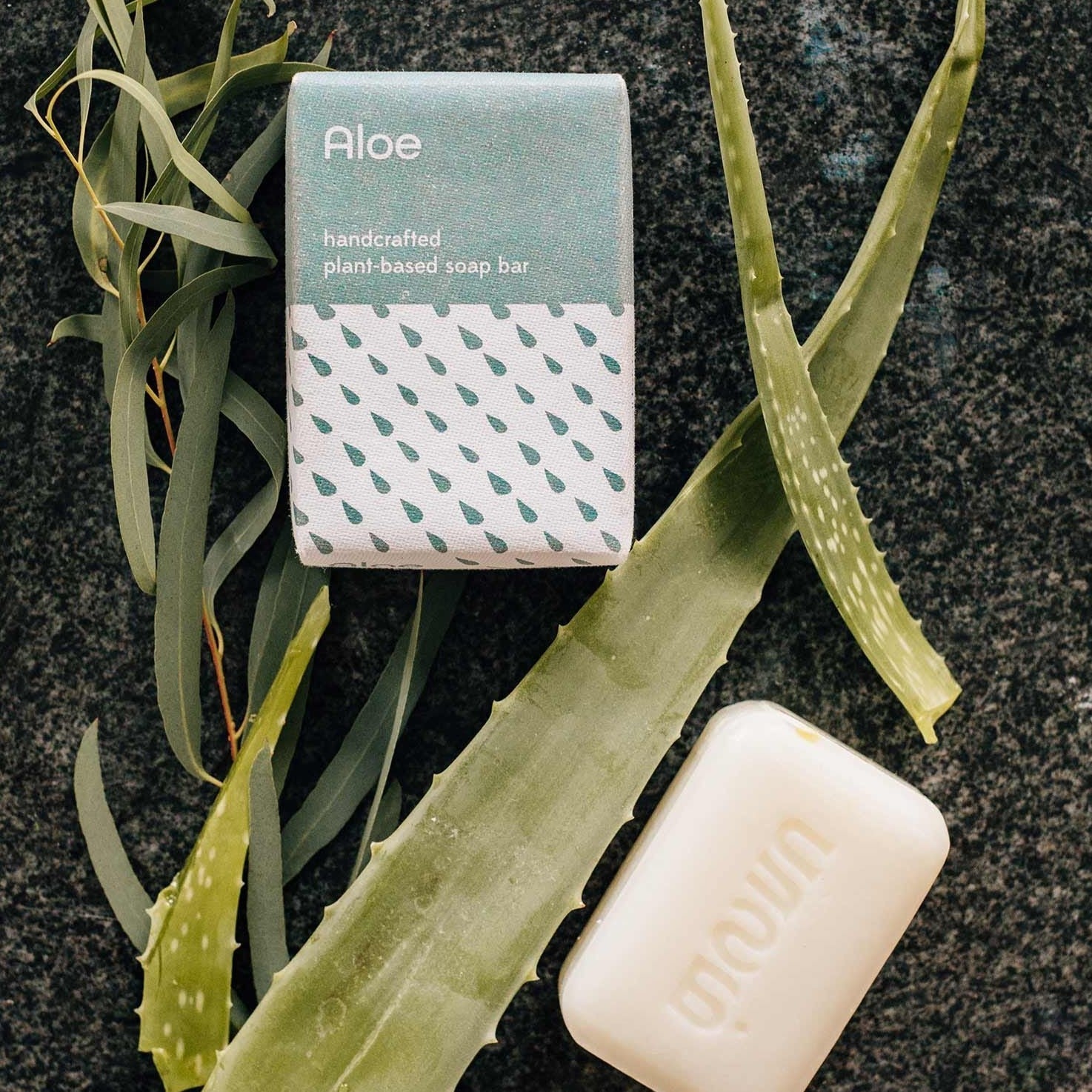 A bar of white aloe scented soap and it's blue and white packaging is placed on a black stone background and shown with aloe leaves.