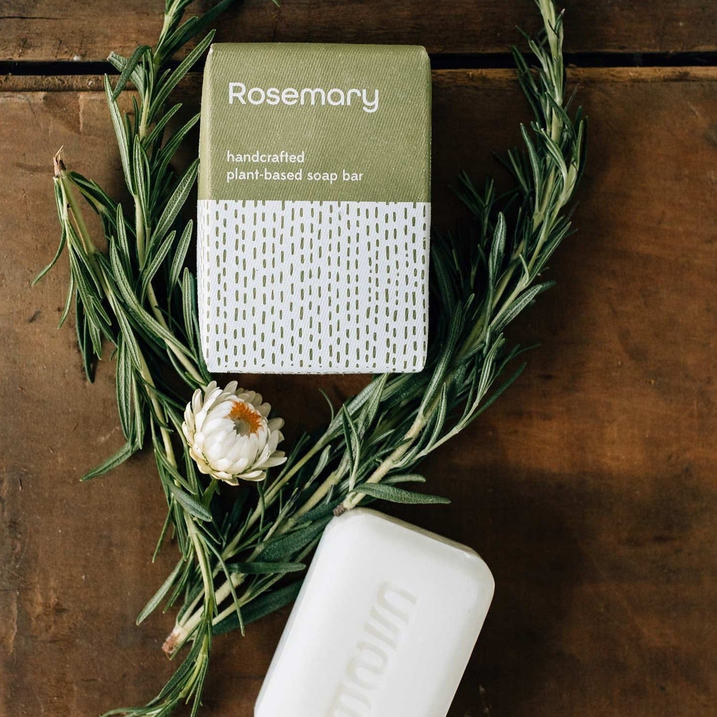 A bar of white soap and its green and white package are placed on a wooden surface and surrounded by rosemary sprigs.