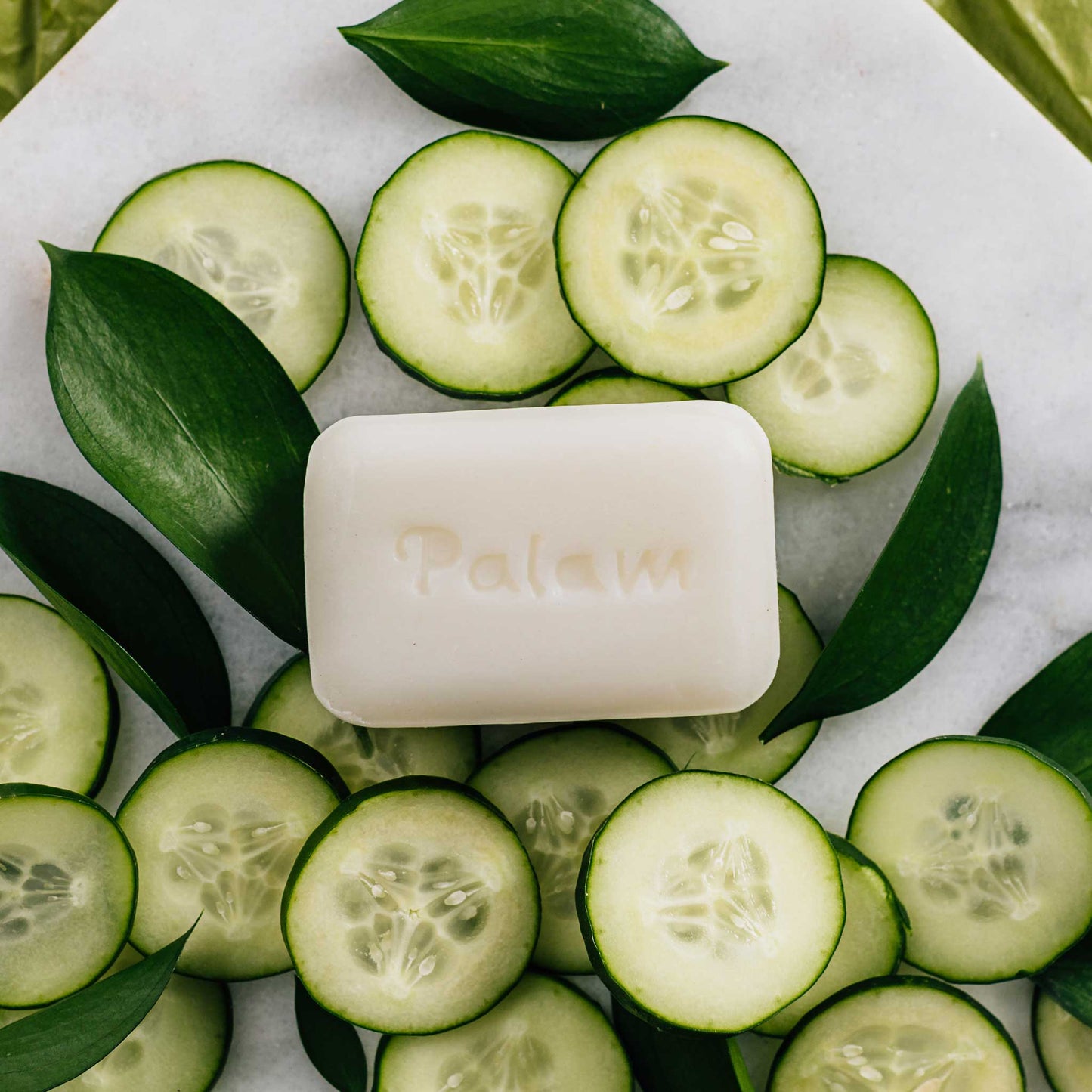 A bar of white soap is laid on top of cucumber slices and leaves on a white stone background.