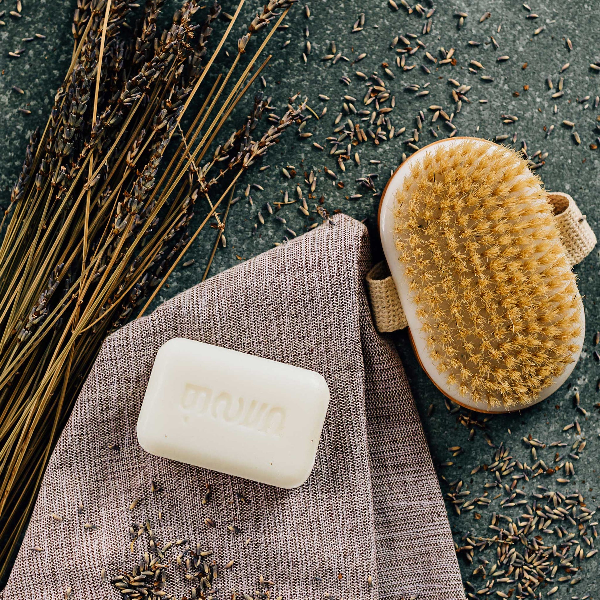 A bar of white soap is styled on a grey cloth and surrounded by a yellow bristle brush, lavender sprigs, and lavender buds.