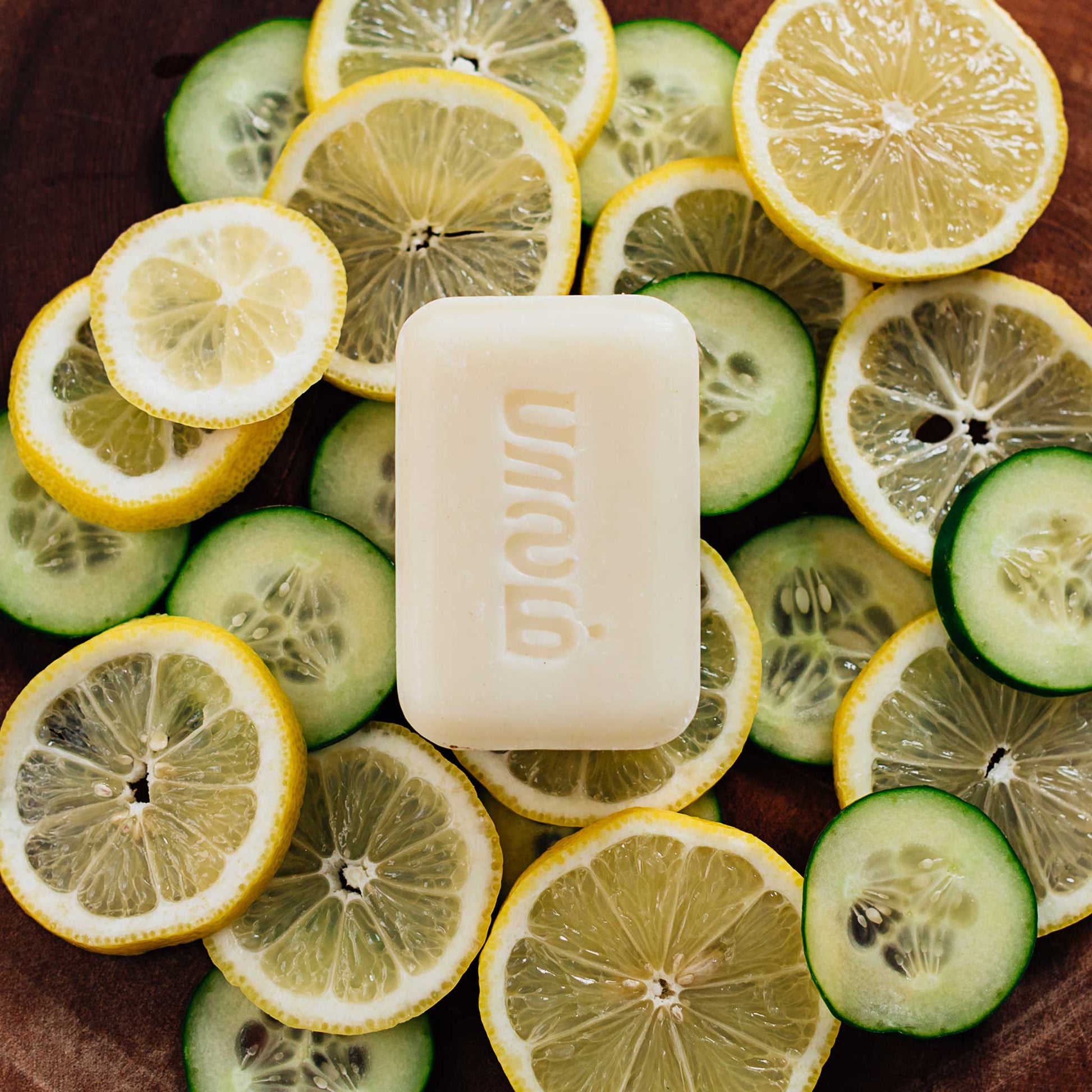 A bar of white soap is laid on top of a bed of lemon and cucumber slices in a wooden bowl.