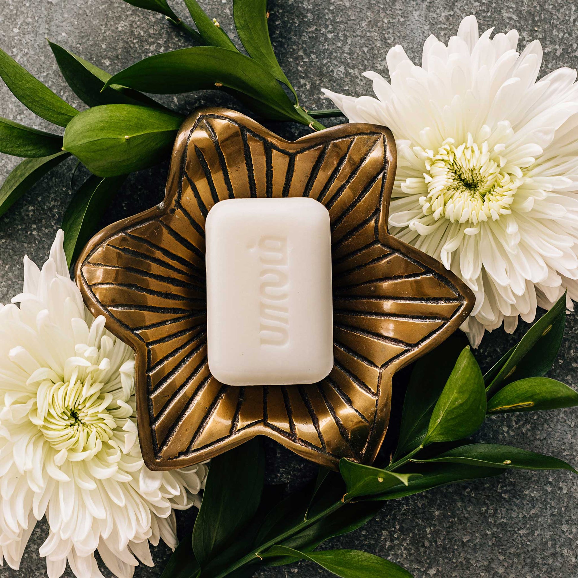 A bar of white soap in a floral brass soap dish on a grey stone surface is surrounded by white flowers.