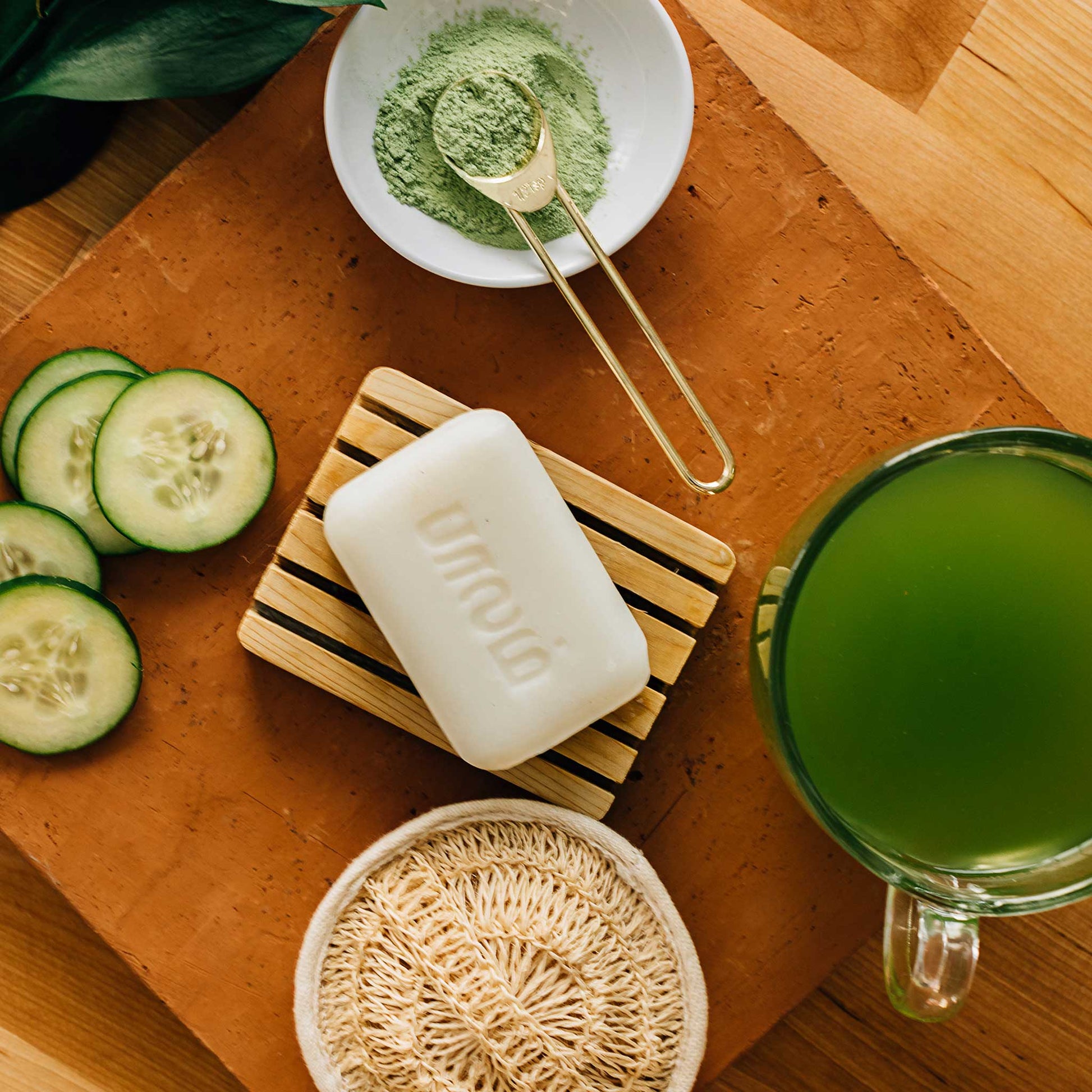 A bar of white soap on a brown slatted soap dish is surrounded by a cup of green tea, green tea powder, cucumber slices, and bath scrubber.