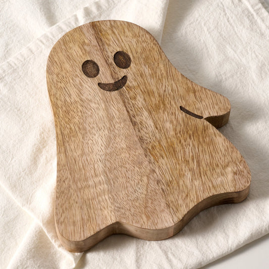 A wooden ghost-shaped serving board with a carved smiling face, resting on a textured off-white fabric background.
