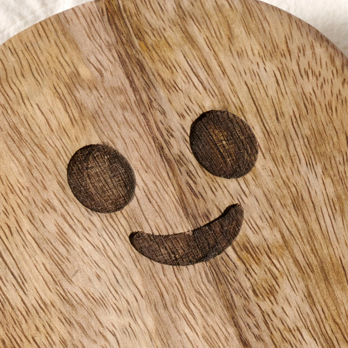 A close-up of a round wooden board or tray with a carved smiley face design, showing the natural wood grain texture.