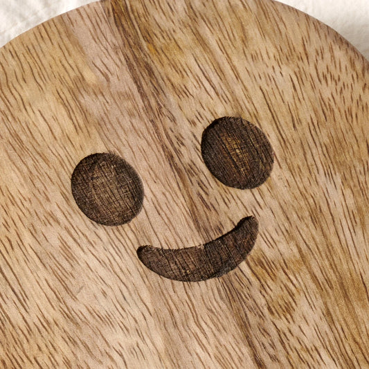 A close-up of a round wooden board or tray with a carved smiley face design, showing the natural wood grain texture.