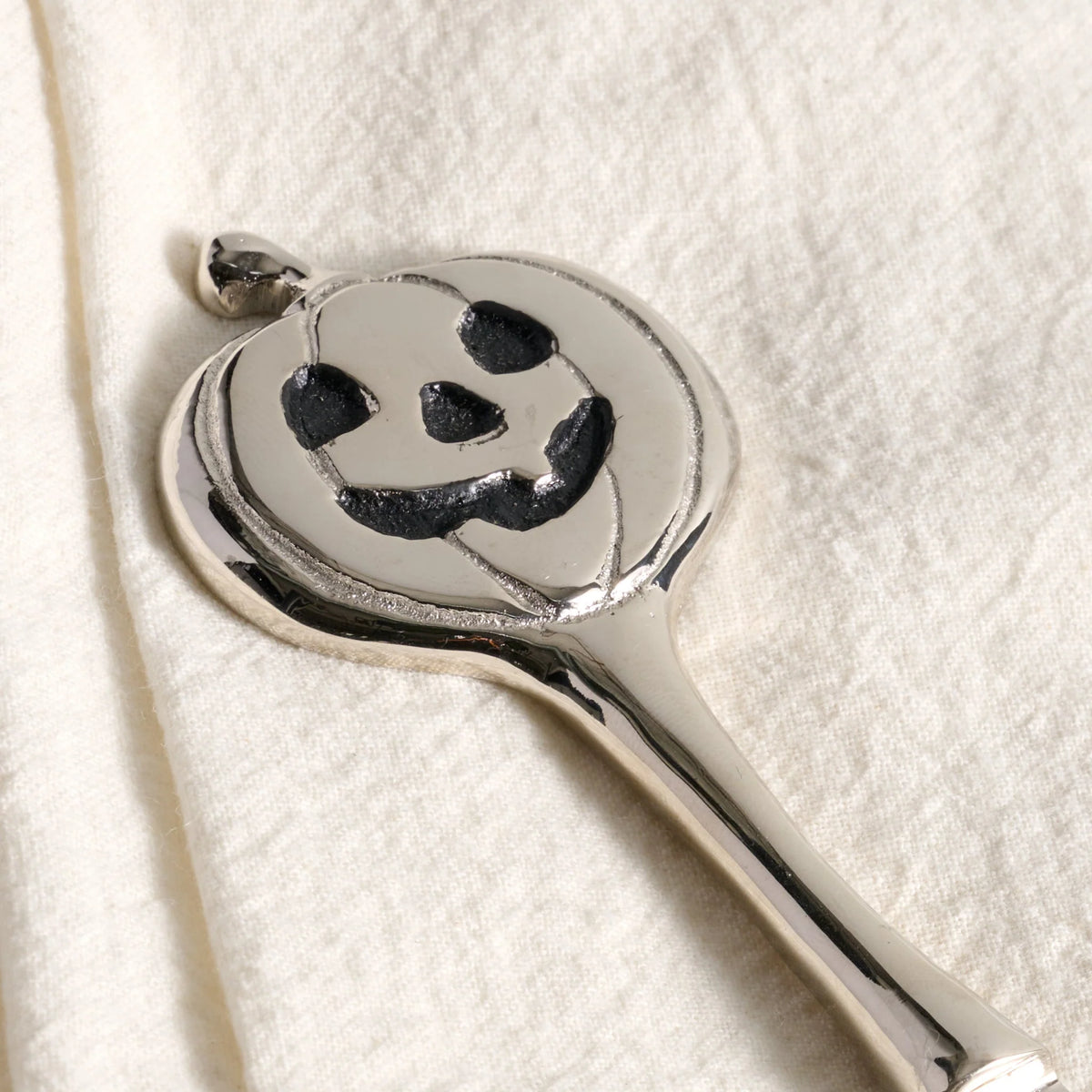A polished silver pumpkin-shaped pendant with a carved jack-o'-lantern face, resting on a textured off-white fabric background.