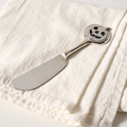 A silver metal cheese knife with a carved jack-o'-lantern handle, resting on a textured off-white linen cloth.