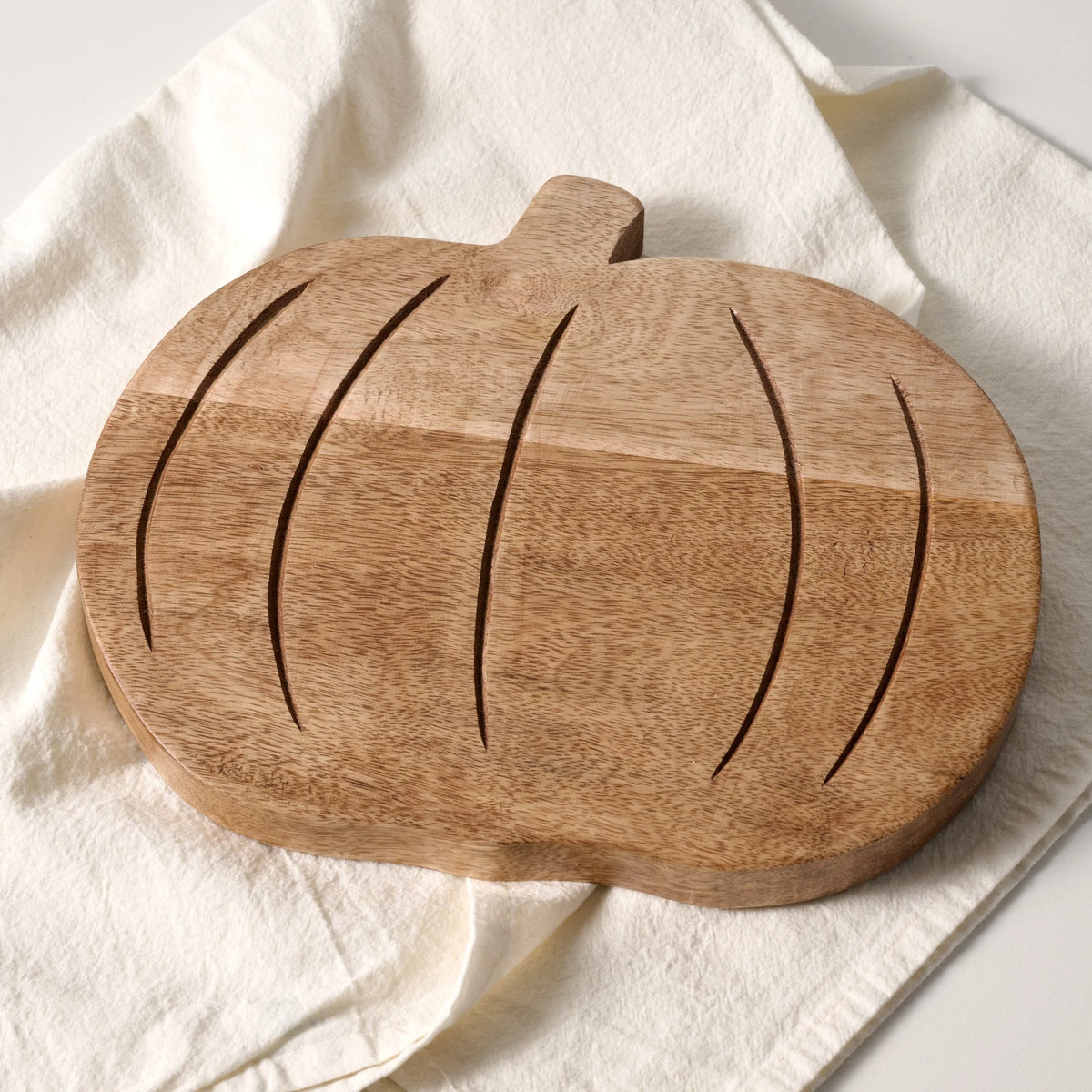 A pumpkin-shaped wooden cutting board with carved vertical grooves and a short stem, photographed on a draped cream fabric background.