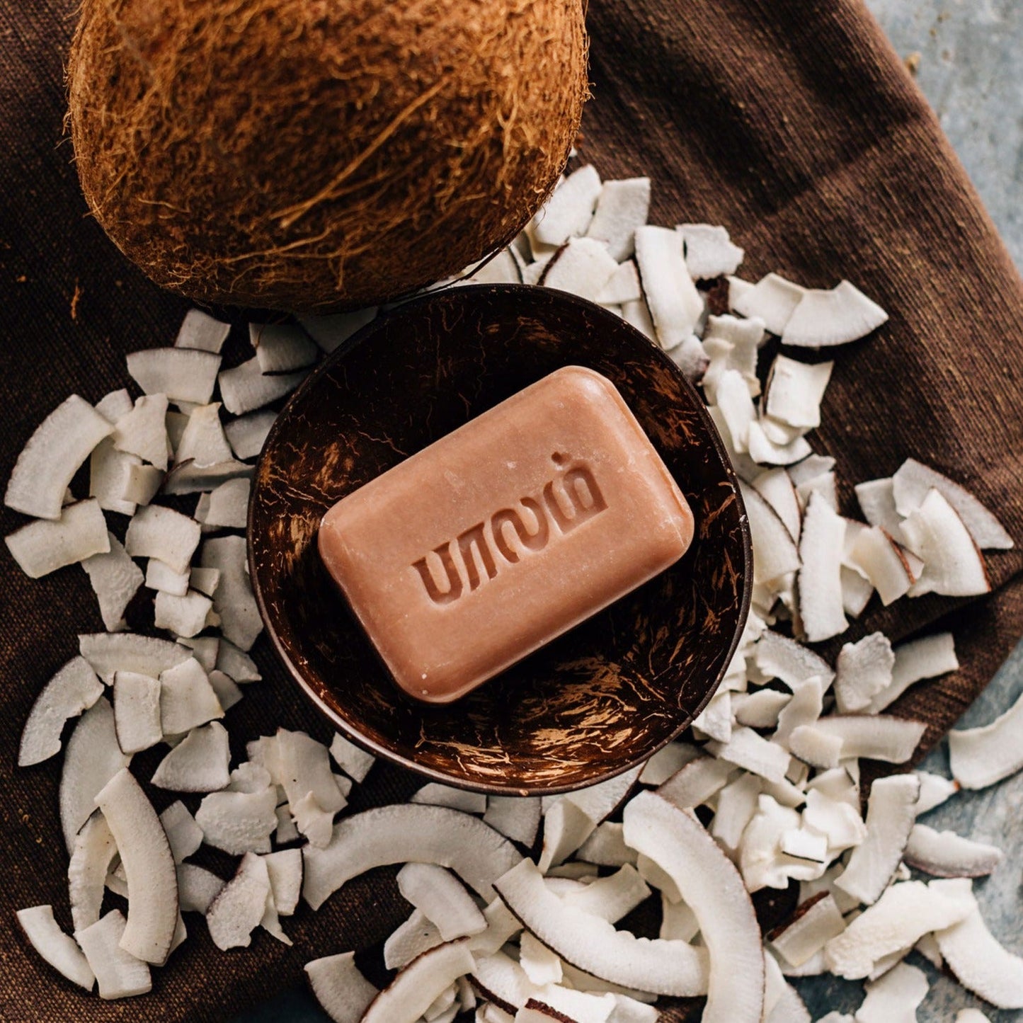A bar of brown coconut soap is set in a coconut shell bowl and surrounded by chips of white coconut flesh.