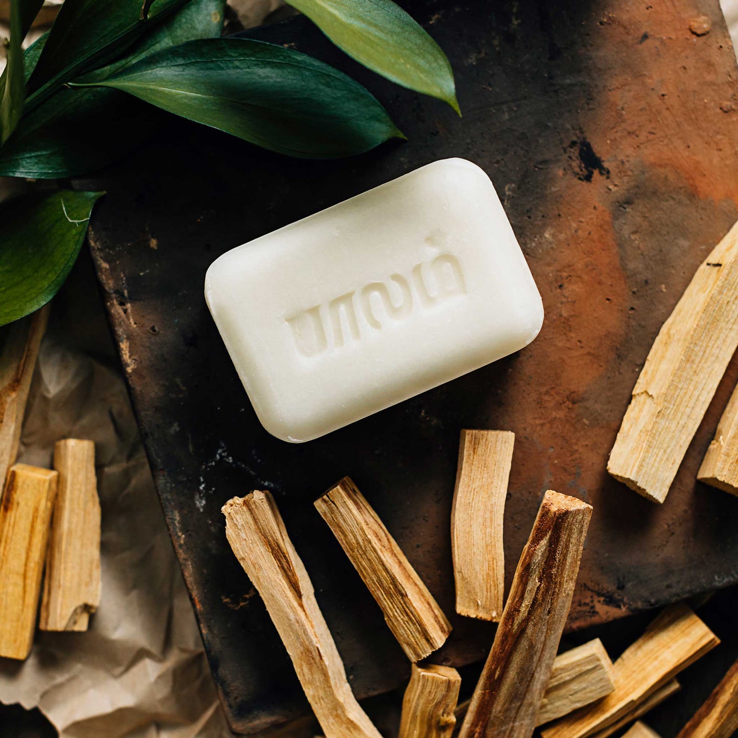 A white bar of soap is laying on a red brick and surrounded by small pieces of brown wood and green leaves.