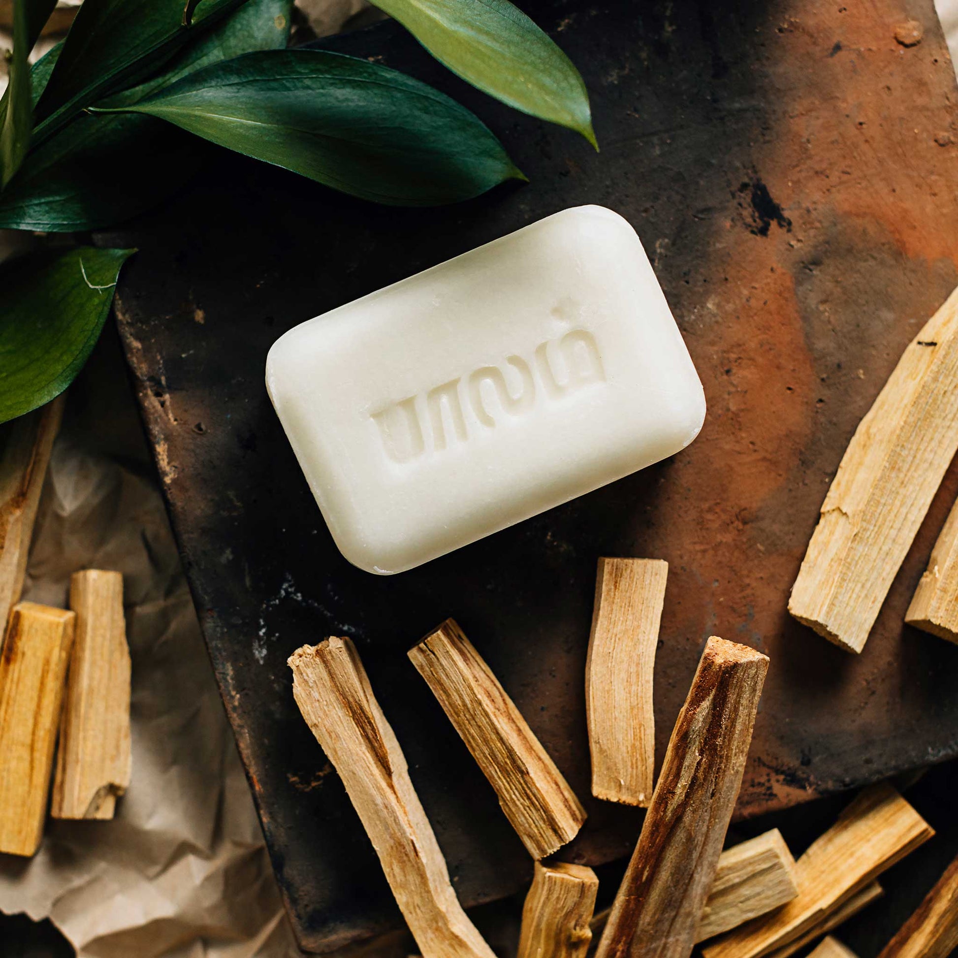 A white bar of soap is laying on a red brick and surrounded by small pieces of brown wood and green leaves.