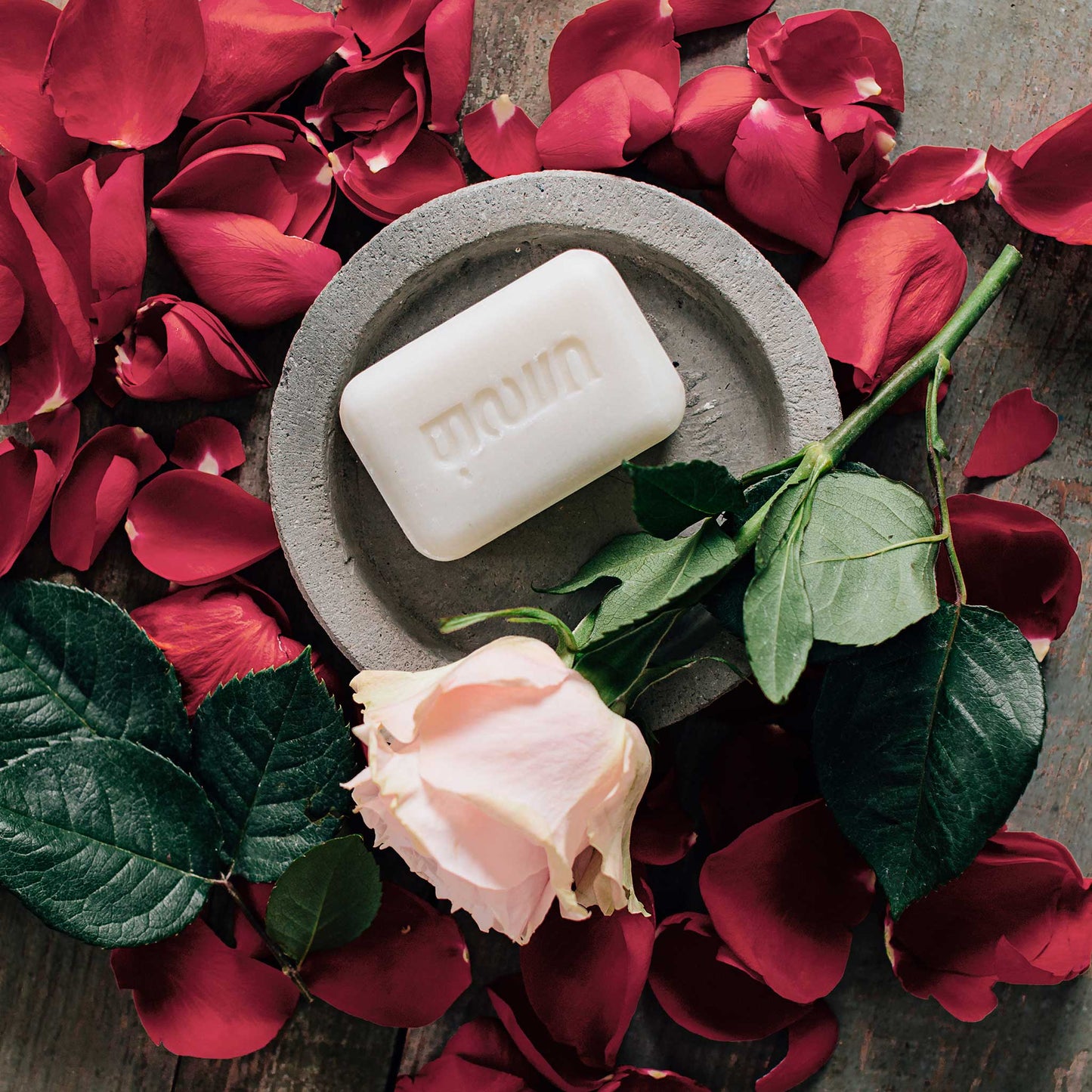A white bar of soap is styled on a grey soap dish and surrounded by red rose petals.