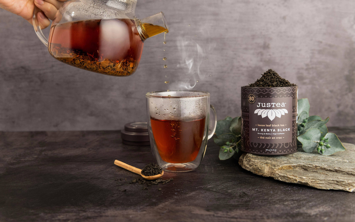 Tea being poured from a glass jar into a mug with a container of Justea tea leaves on a dark surface.