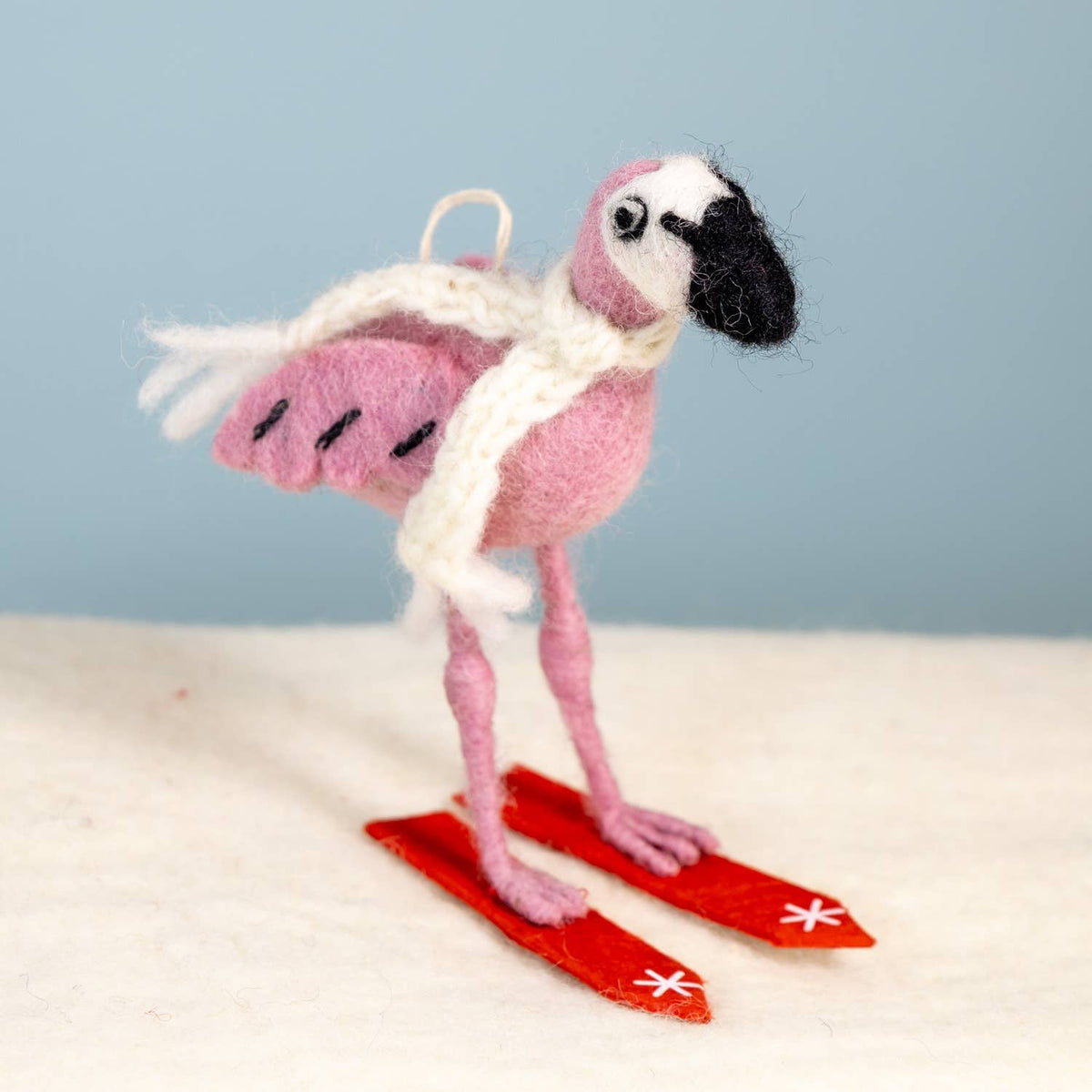 A pink felted wool flamingo ornament wearing a white knit scarf and red felted wool skis, standing on a textured white surface against a light blue background.