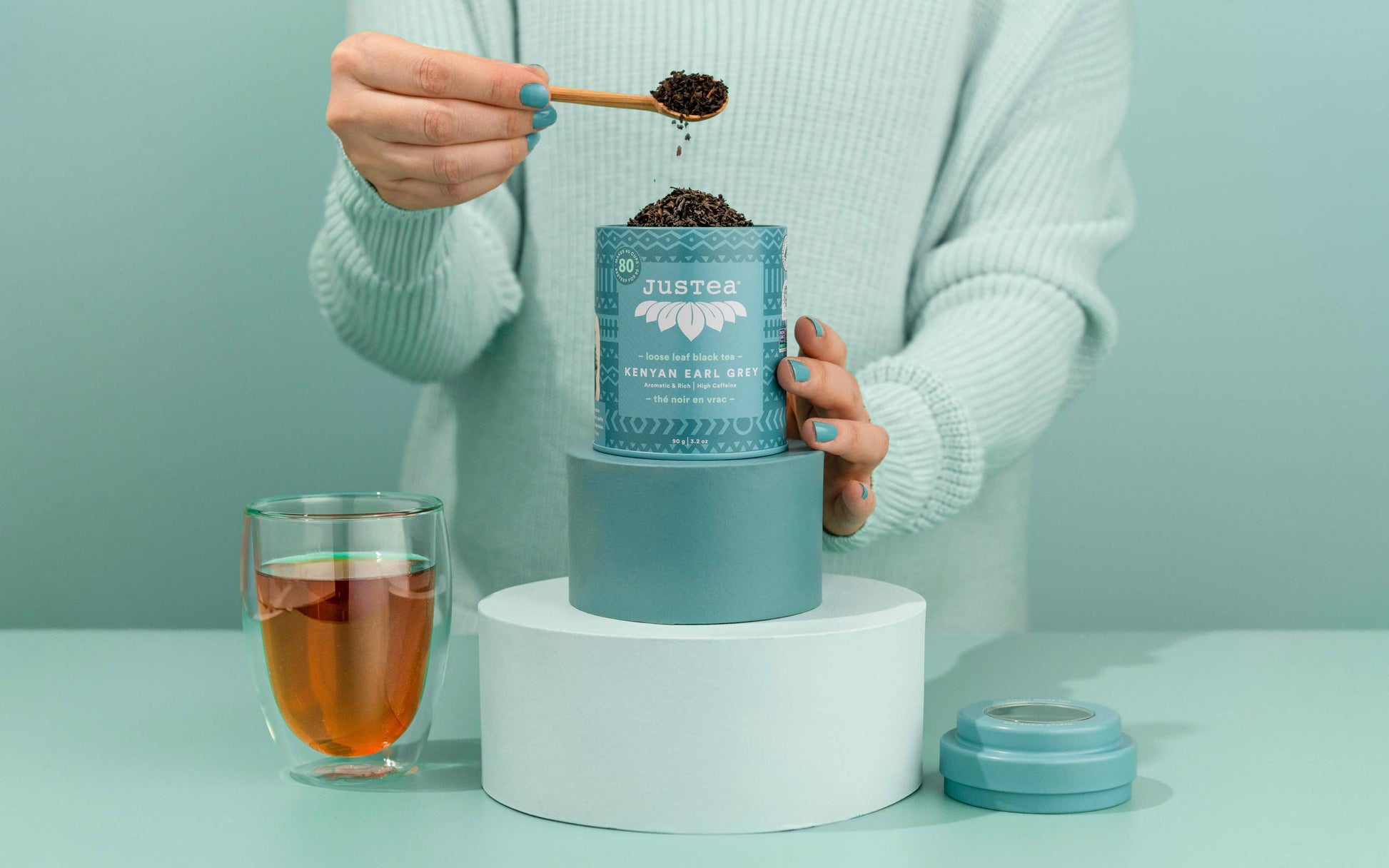 Person using a small spoon to scoop black tea leaves into a teal tea tin labeled JUSTEA on a light green background.
